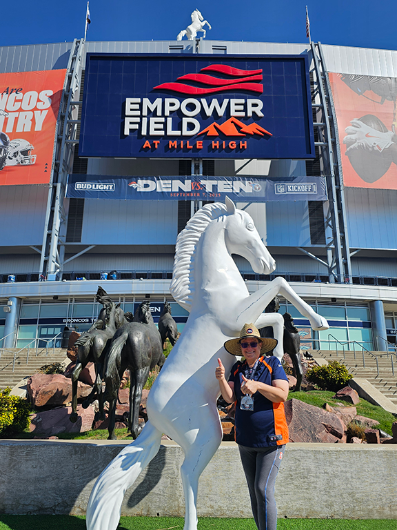 Person in Denver Broncos attire giving thumbs-up beside a white rearing horse statue at Empower Field at Mile High